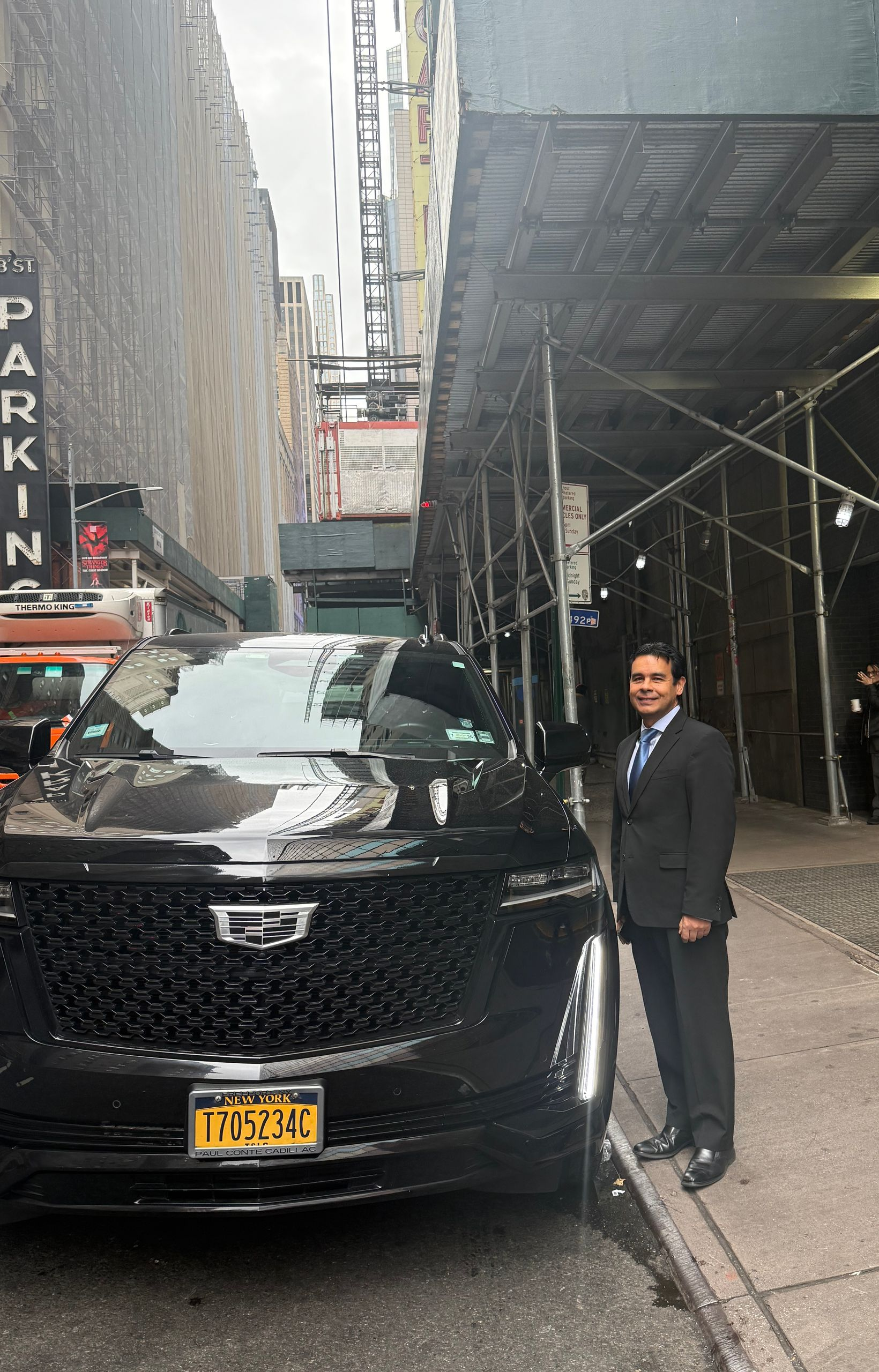 Man in suit next to black SUV, New York City street, parked near a building and scaffolding.