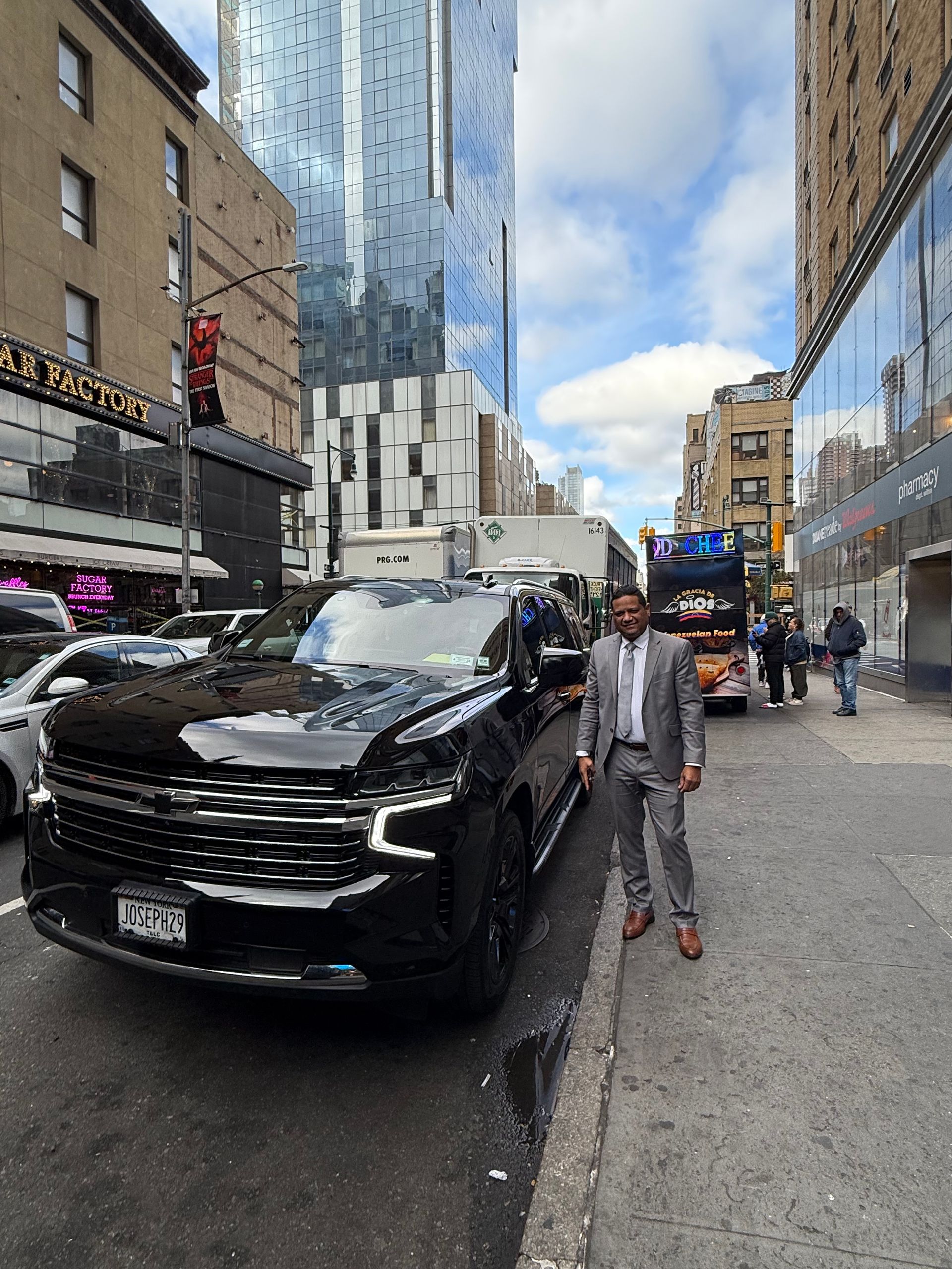 Man in a suit stands next to a black SUV on a city street with tall buildings.