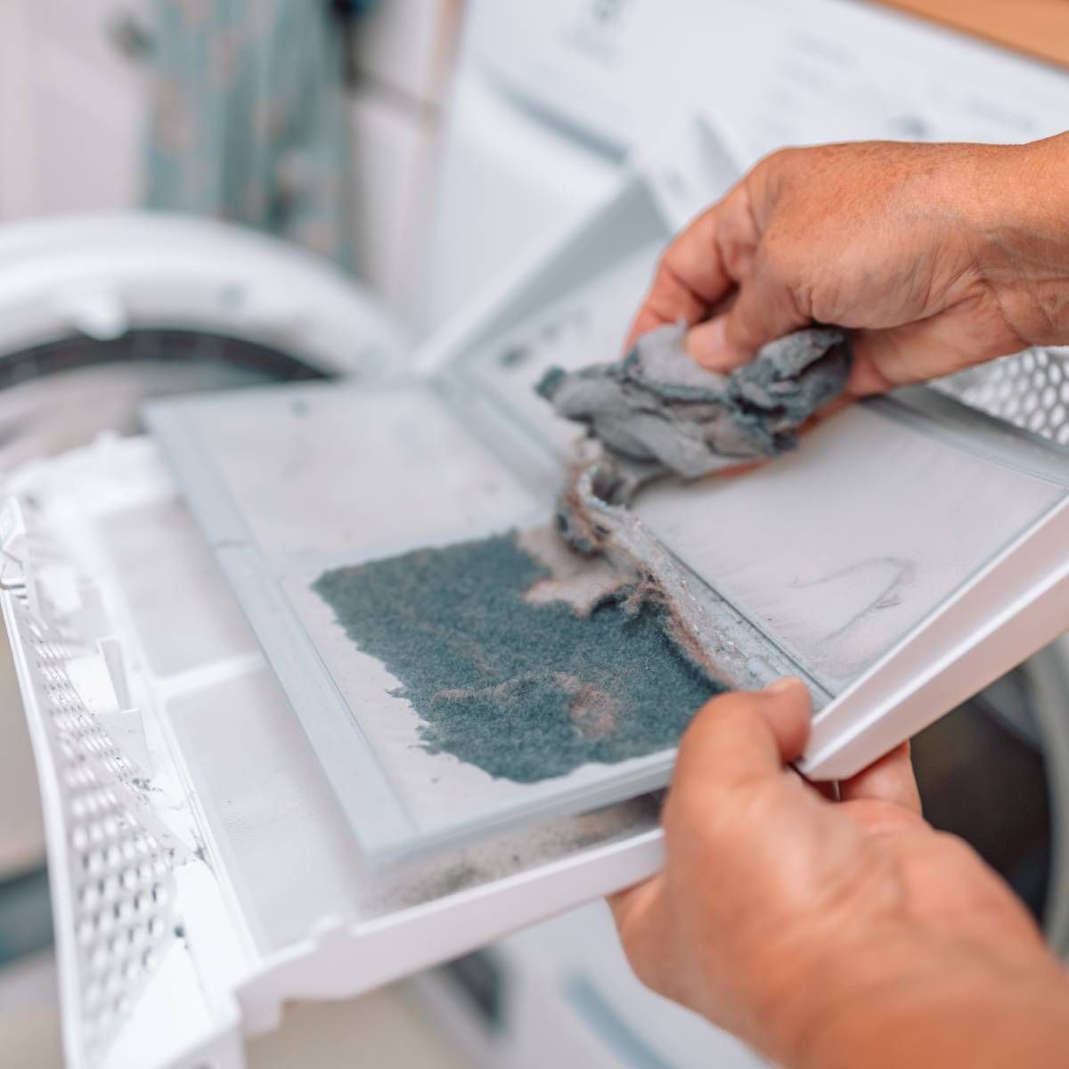 Hands cleaning a dryer lint trap, removing gray lint from a white screen in a laundry room setting.