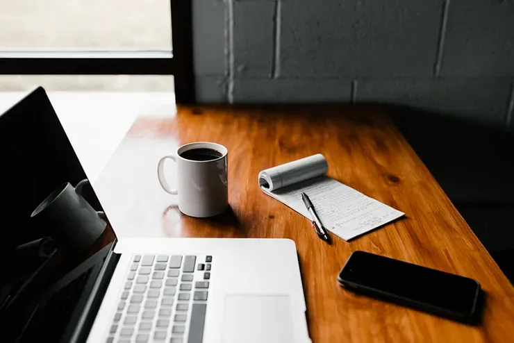 Una scrivania di legno con un computer portatile, una tazza di caffè, un quaderno e un telefono cellulare.