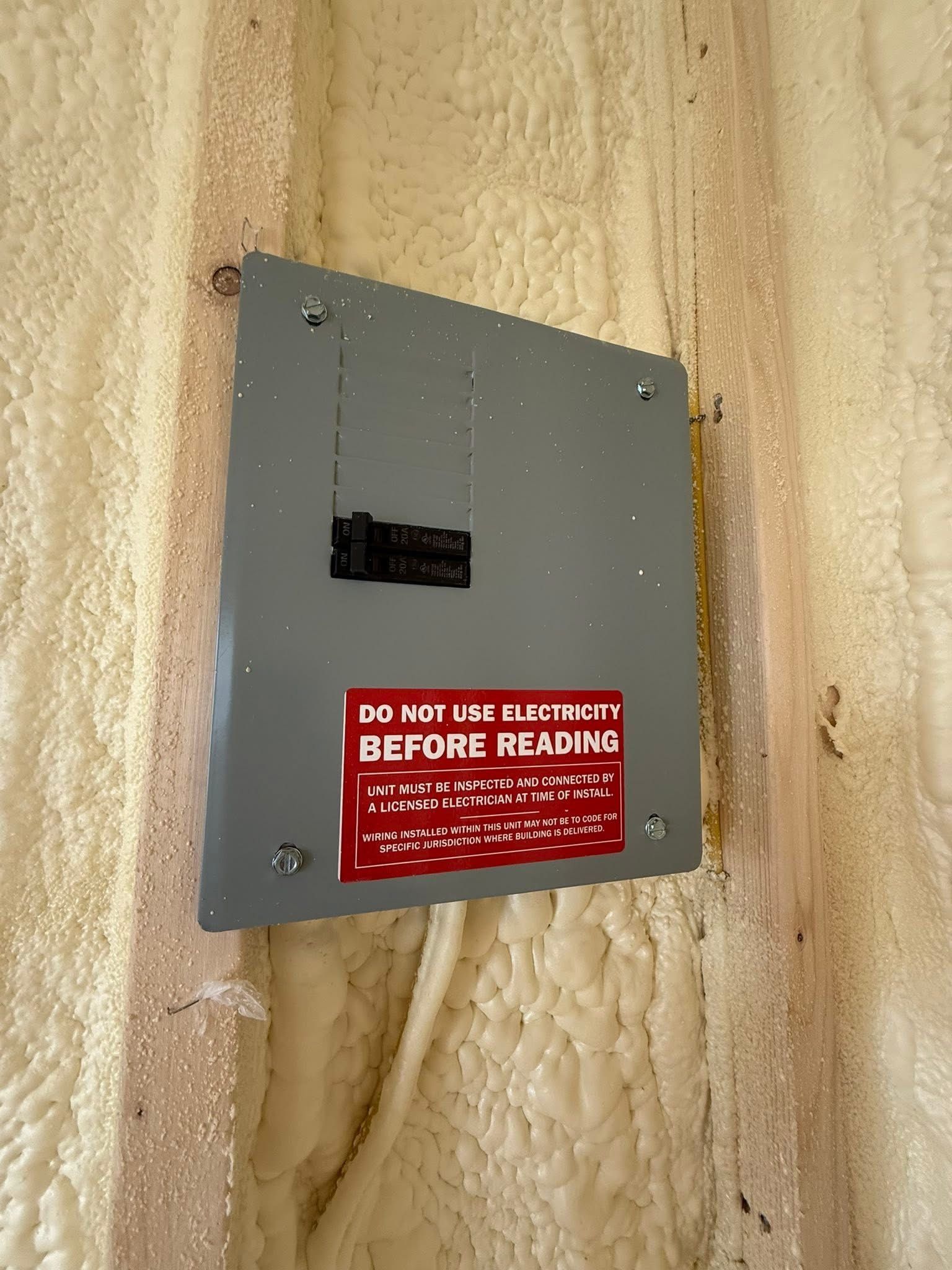 A gray electrical panel with a red warning label is mounted on a wall with beige spray foam insulation.