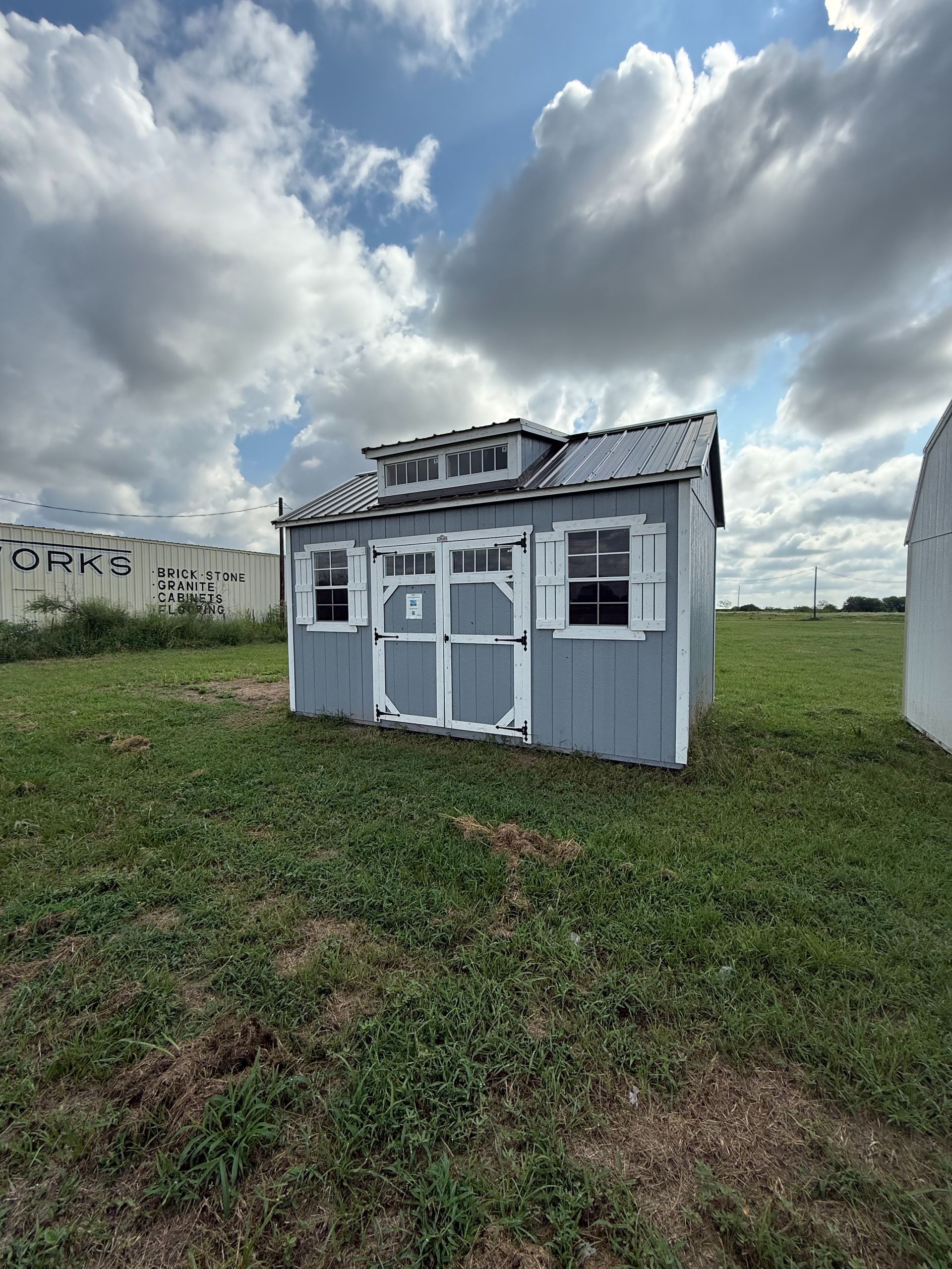 Blue and white shed with a small cupola and shutters, on a grassy field under a cloudy sky.