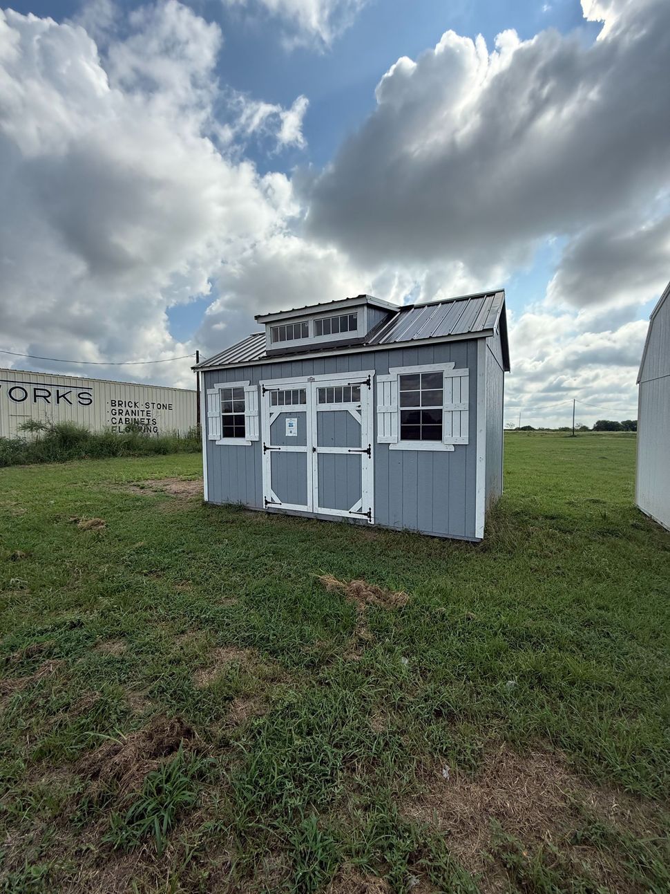 Blue shed with white trim and shutters, sitting in a grassy field under a cloudy sky.