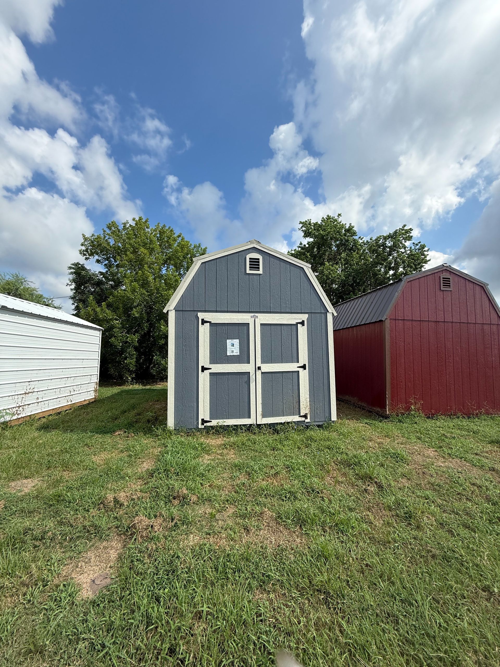 Blue shed with white trim and double doors on a grassy lot, under a blue sky with clouds.