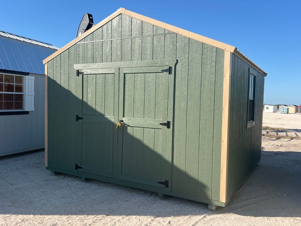 Green wooden shed with beige trim and double doors on a gravel lot under a clear sky.