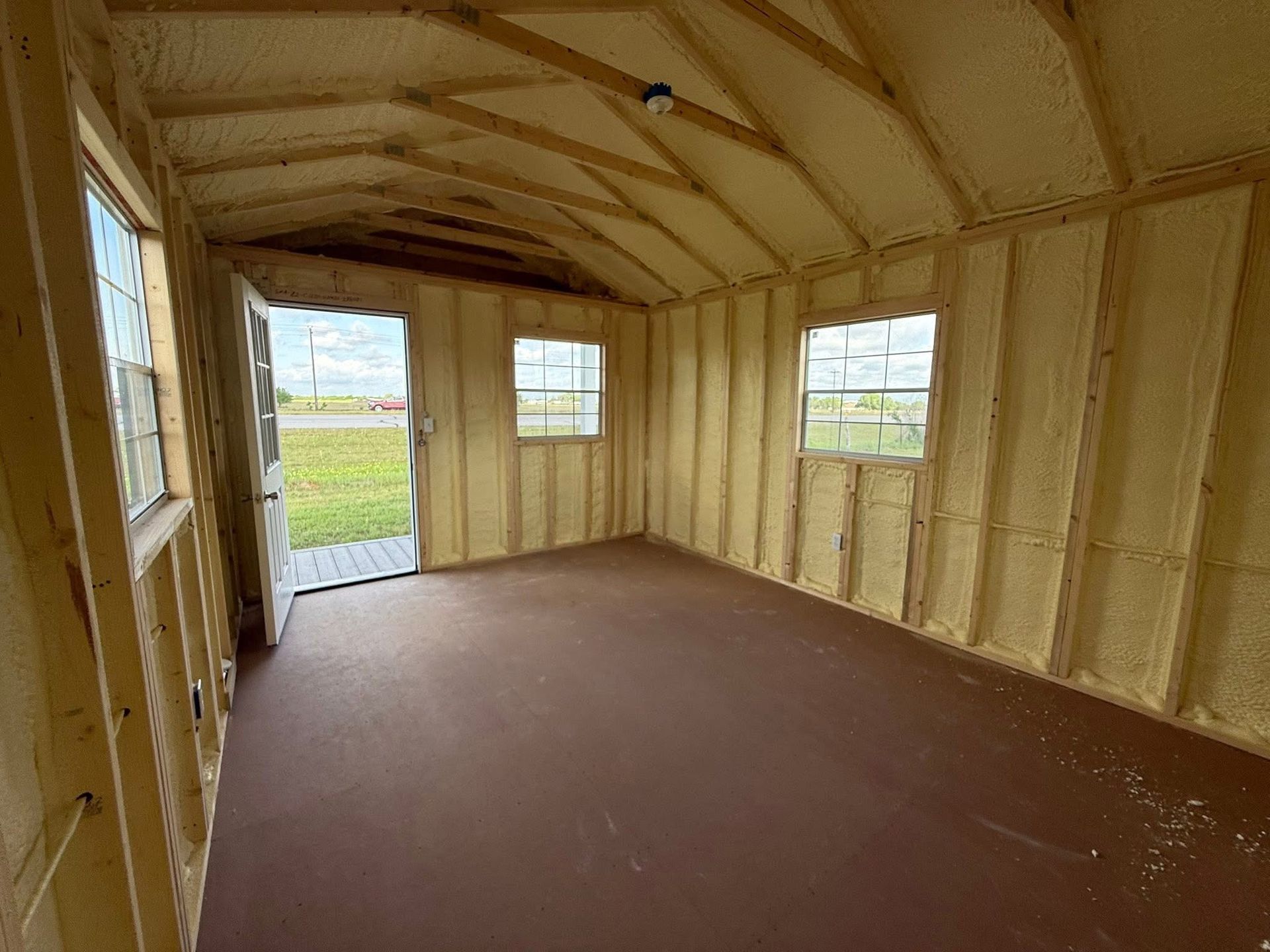 Interior of a small cabin with spray foam insulation visible, open door, windows, and brown floor.