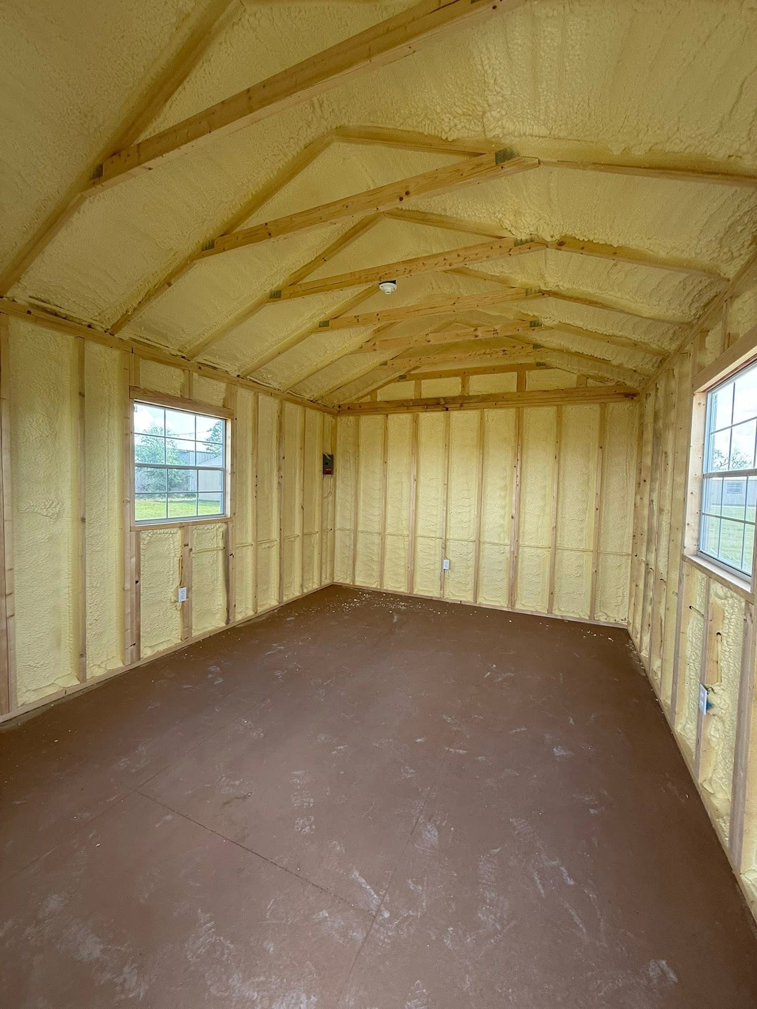 Interior view of a wooden shed with spray foam insulation, two windows, and a brown floor.