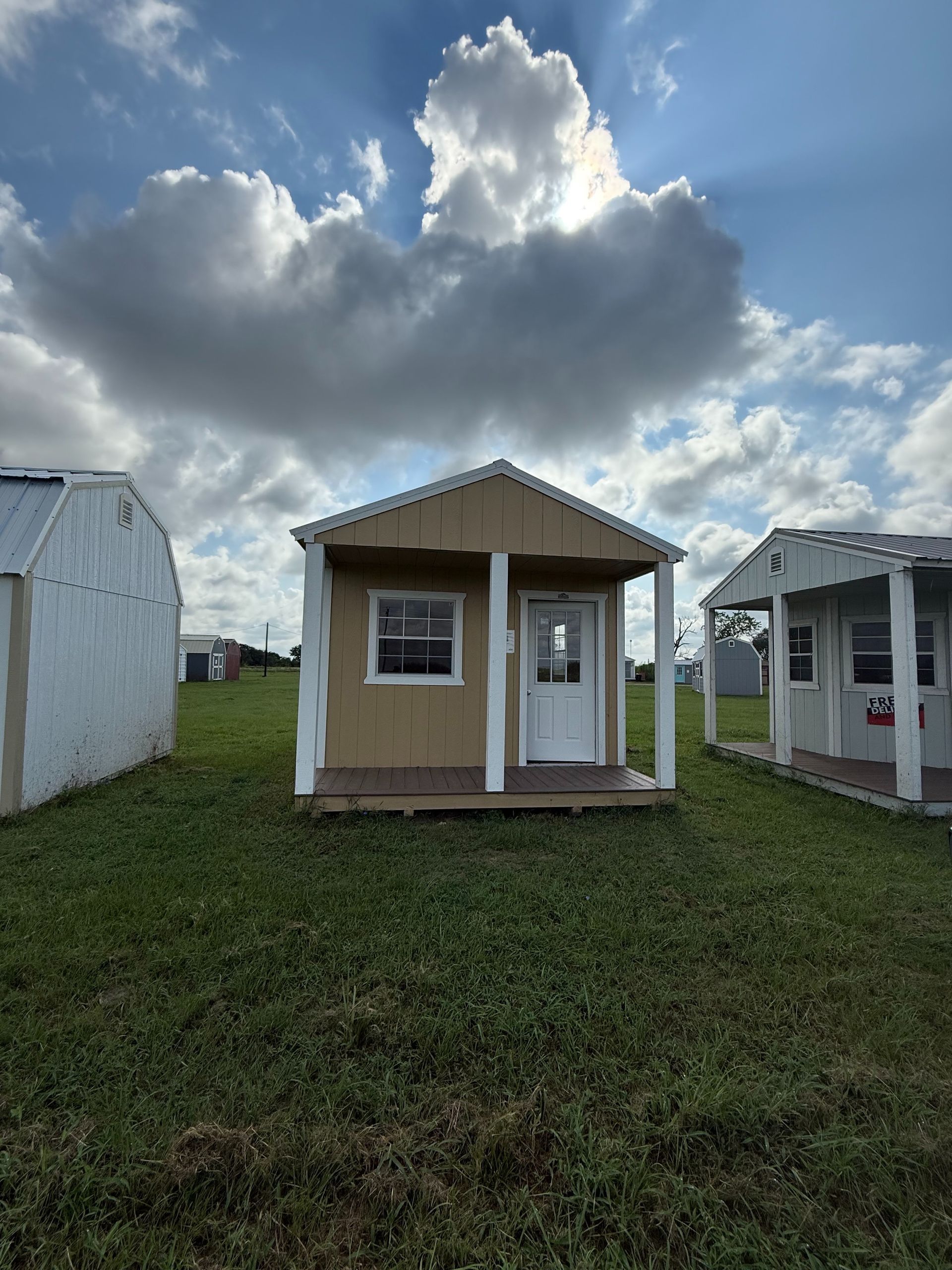 Small tan shed with white trim and a front porch, on a grassy field, under a cloudy sky.