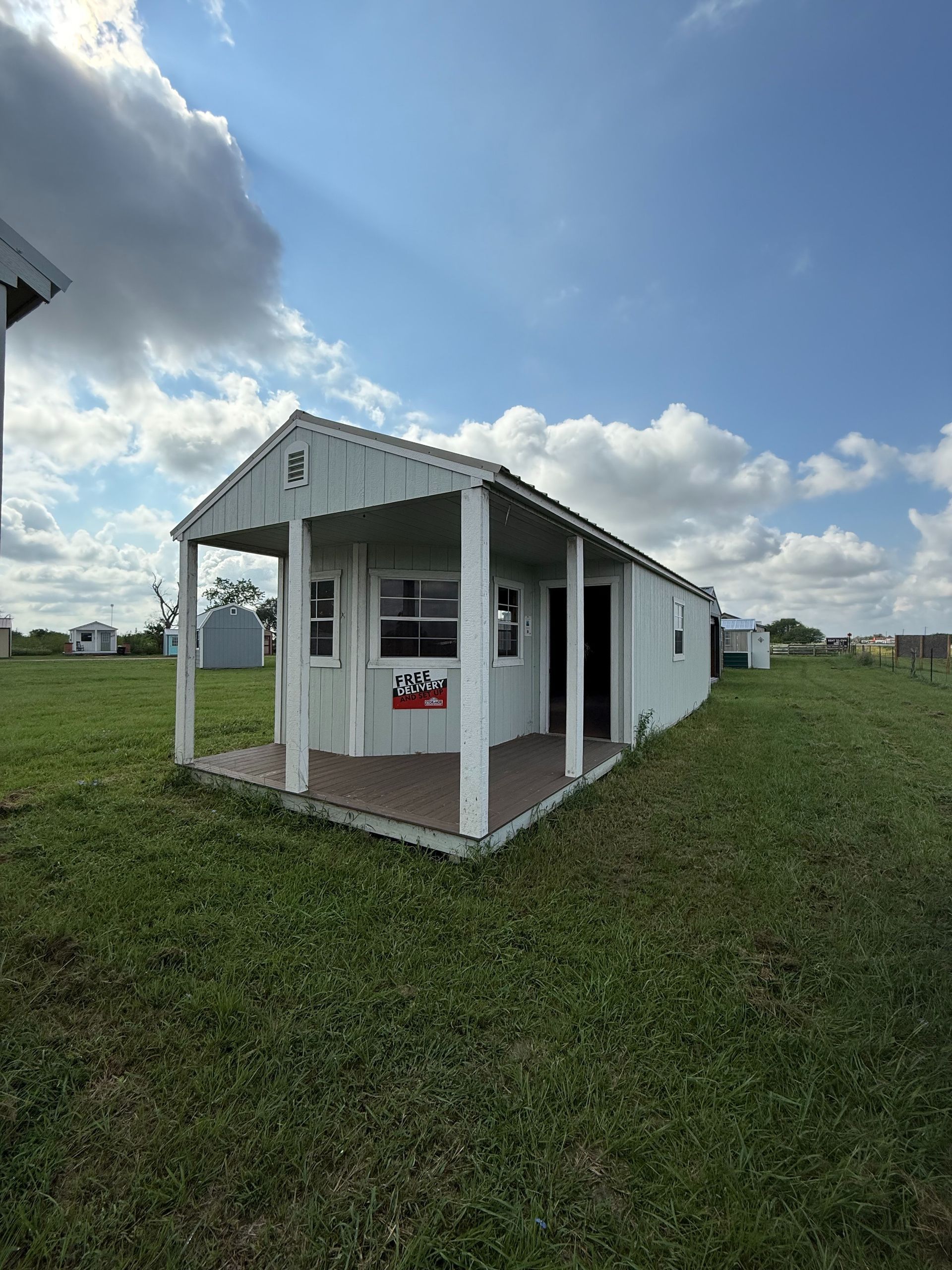 White cabin with a porch on a grassy field under a partly cloudy sky.