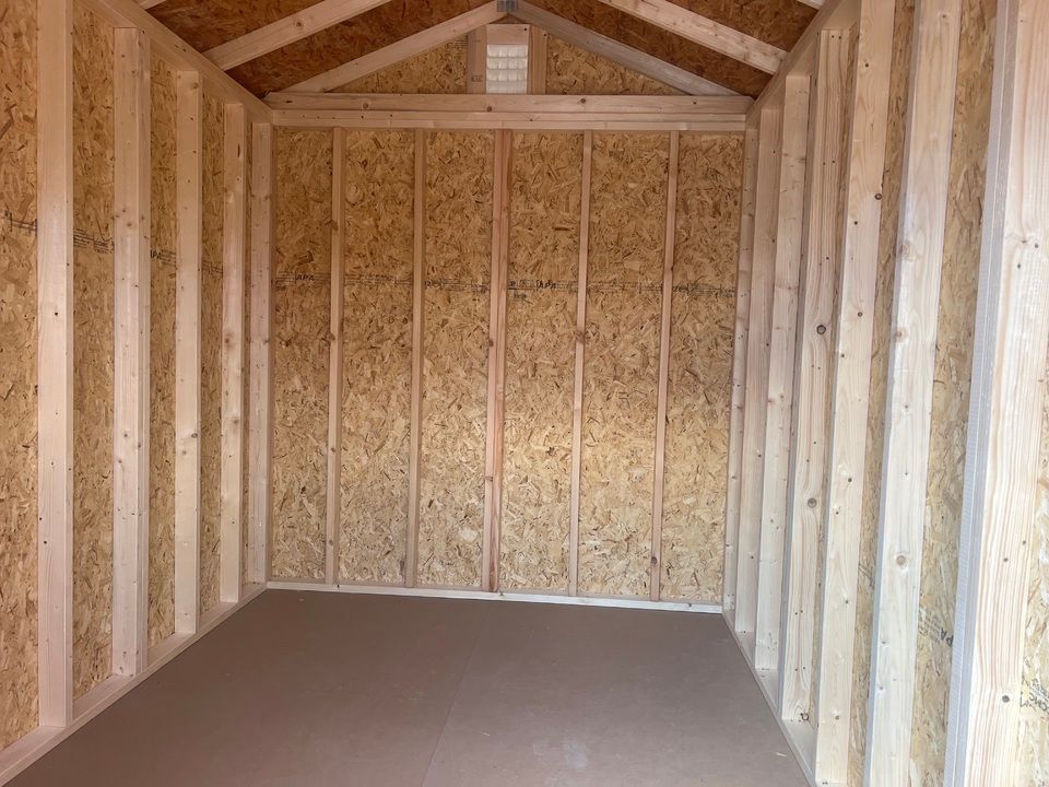 Interior view of an empty wooden shed, with walls and roof made of light-colored wood. The floor is covered in brown material.