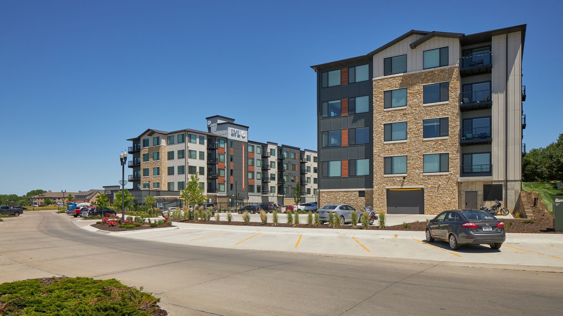 Apartment complex on a bright day; buildings with stone and dark siding, cars parked on the street.