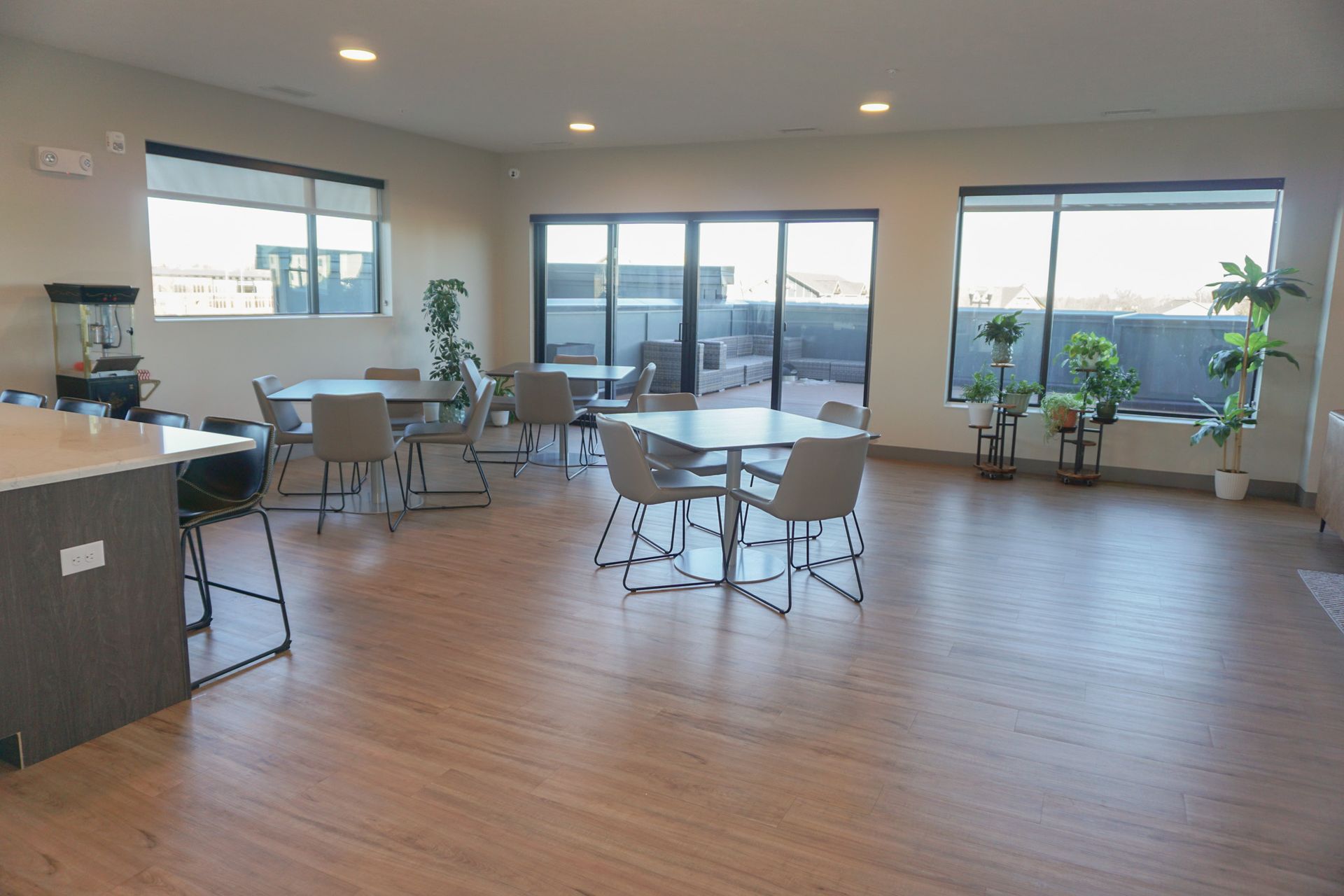 Spacious community room with tables and chairs, large windows, and plants. Light wood floors and neutral walls.