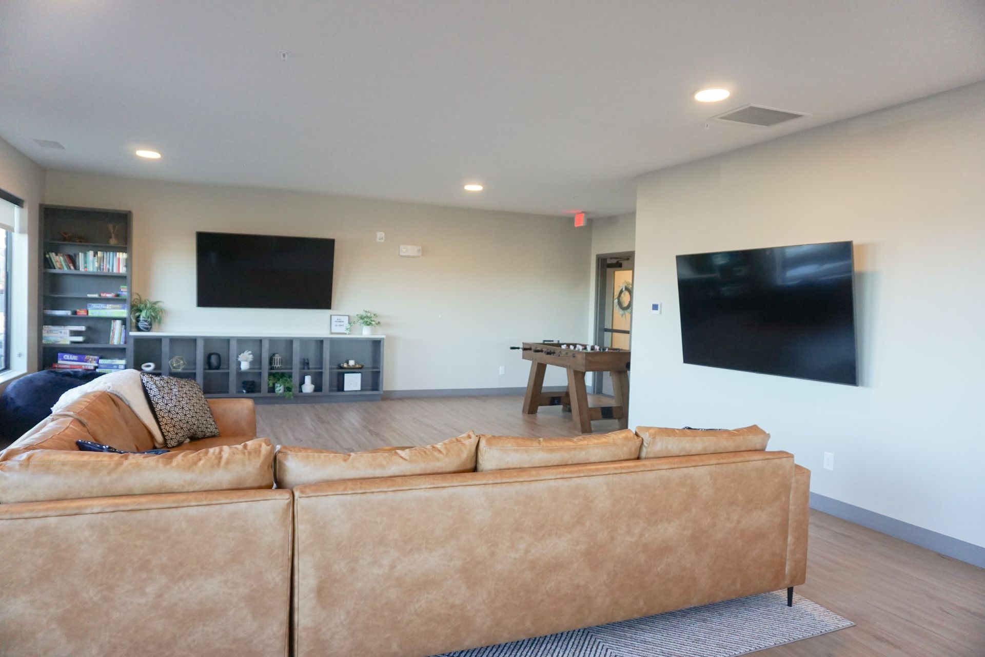 Spacious living area with tan sectional sofa, two TVs, a foosball table, and shelving.