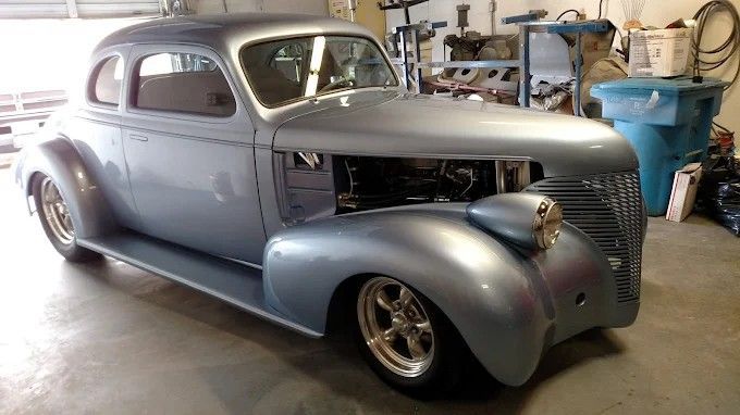A silver vintage coupe parked inside a garage, featuring a rounded hood, prominent grille, and chrome custom wheels.