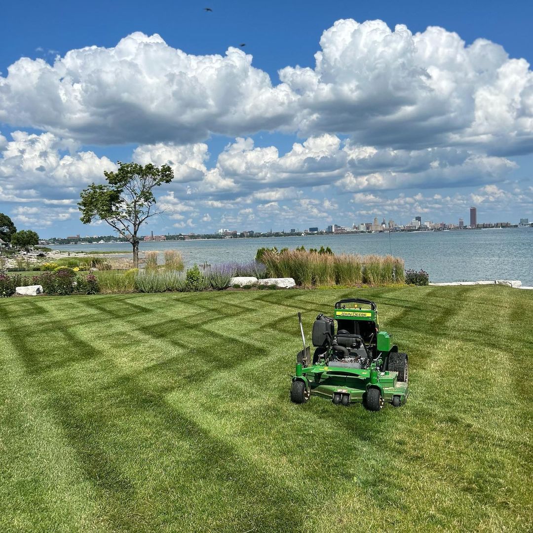 A green lawn mower is cutting a lush green lawn next to a body of water.