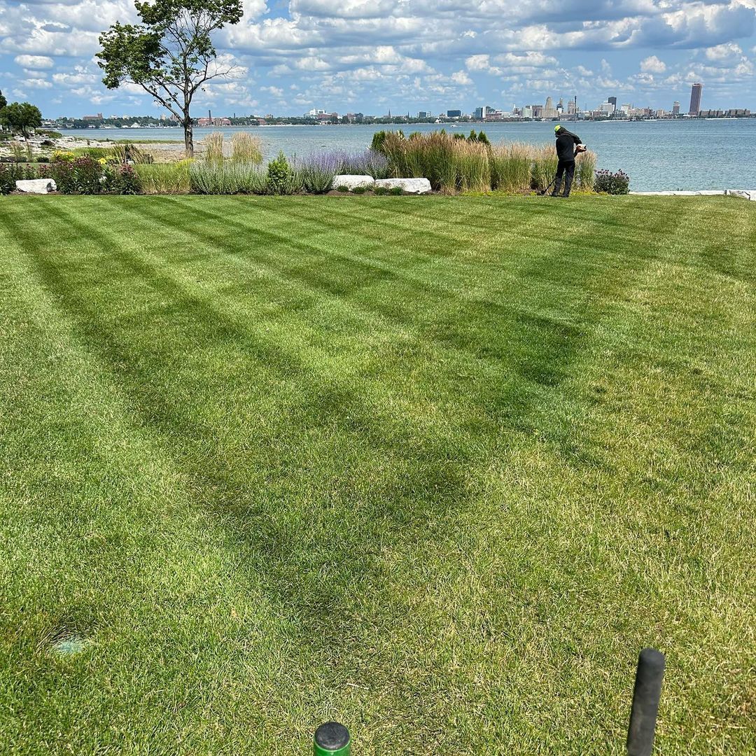 A man is mowing a lush green lawn next to a body of water.