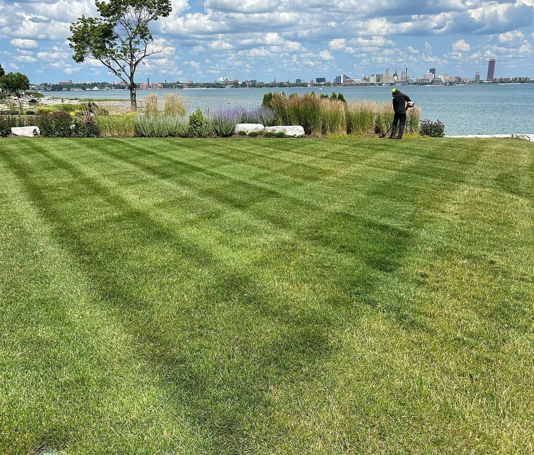 Lawn mowed with stripes, person working, lake and city skyline in background, under a cloudy sky.