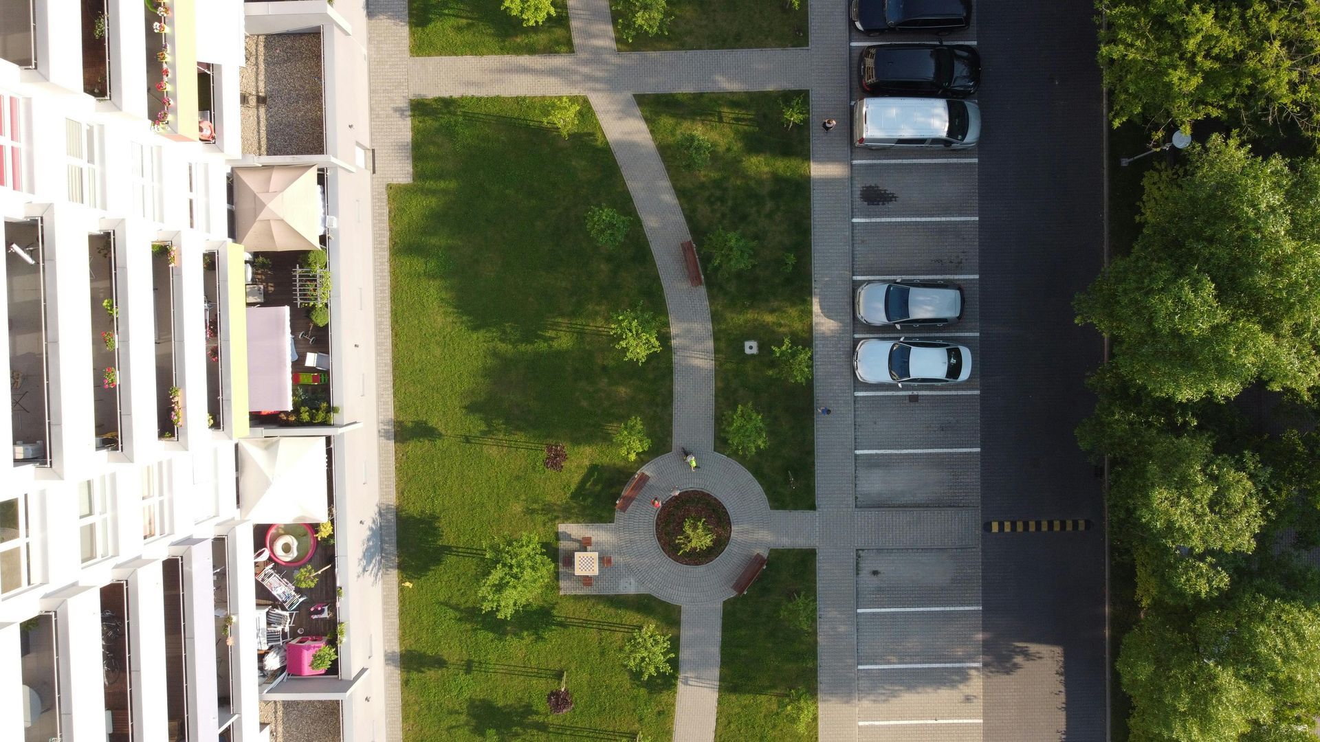 Overhead view of a building, green space with a path, and a parking lot with parked cars.