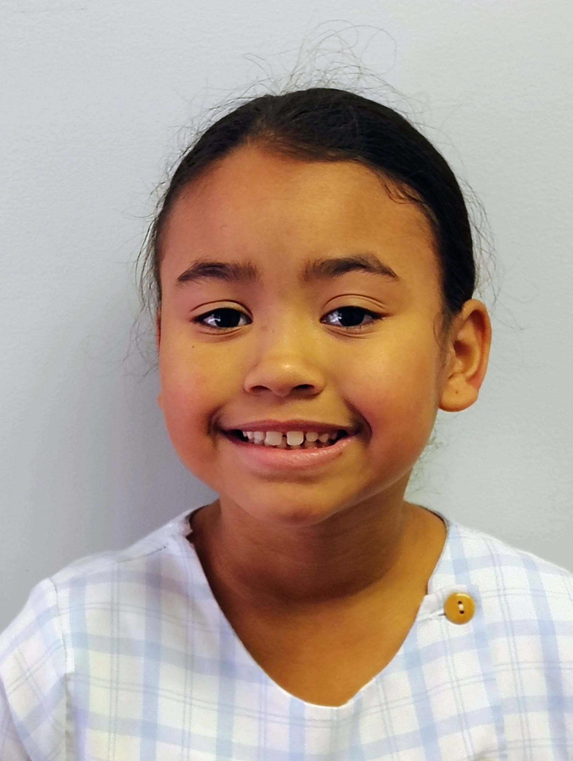 Young girl smiling at the camera, wearing a light blue checked top with a brown button.