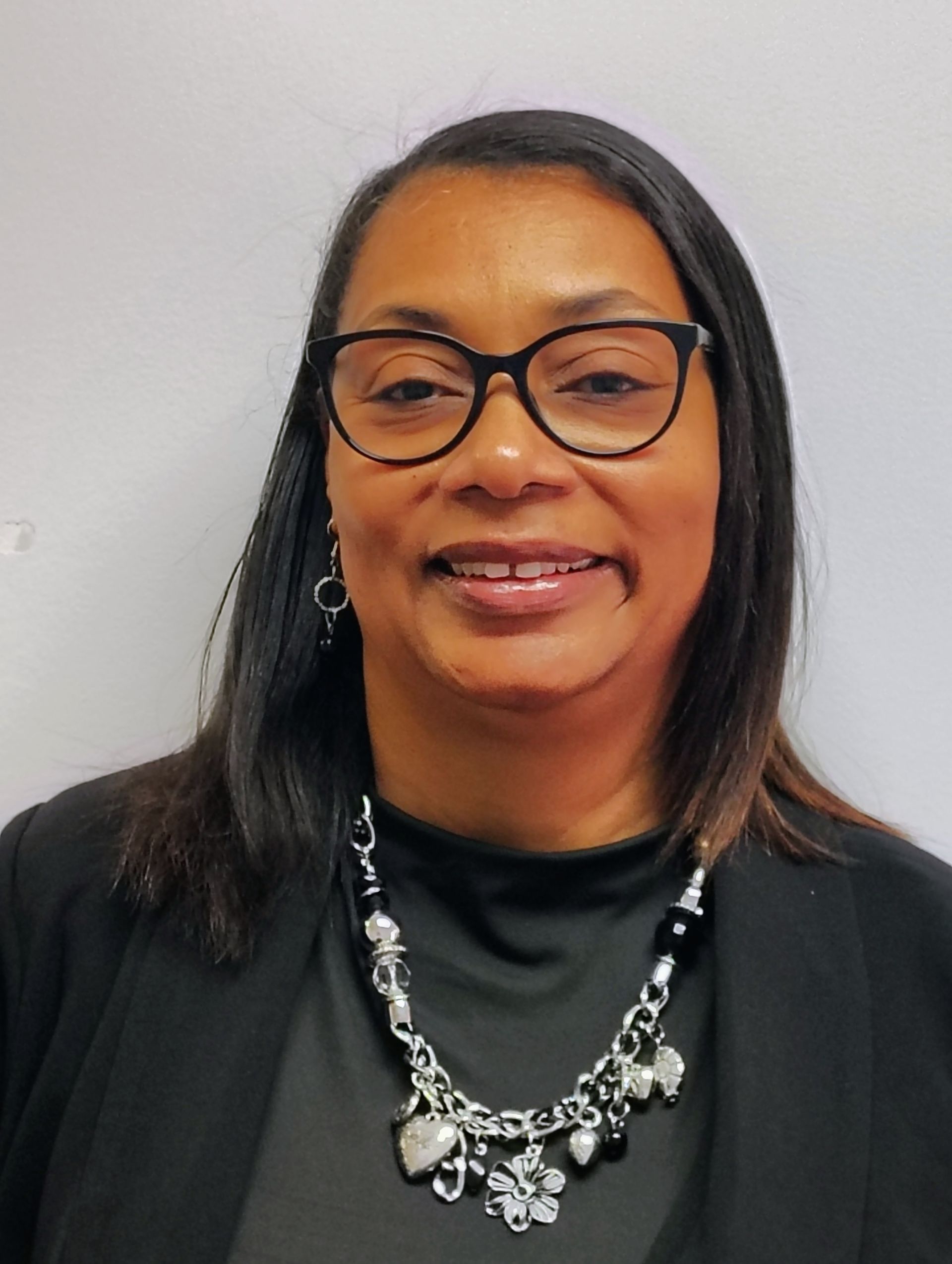 Woman with glasses, wearing a black blazer and necklace, smiling in front of a white background.