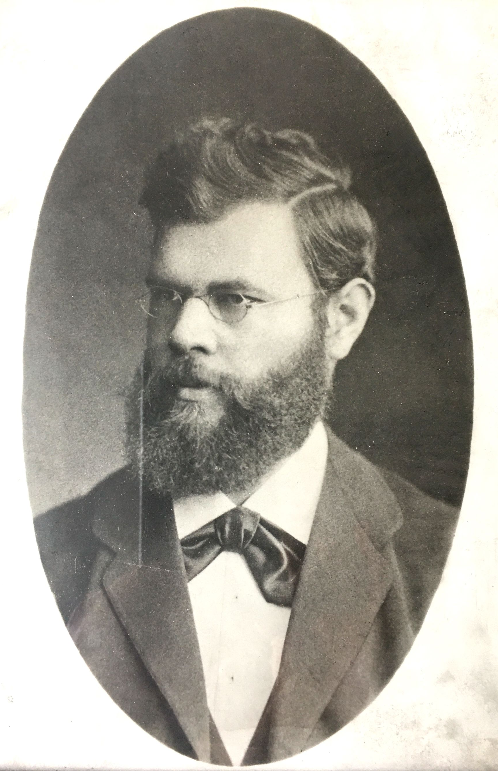 Mann mit Brille und Bart, der eine Fliege und ein Sakko trägt, Porträt in einem ovalen Rahmen.