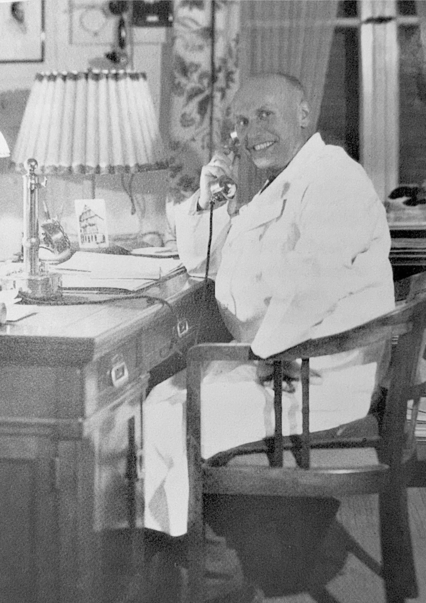 Ein Mann in weißer Kleidung sitzt lächelnd an einem Schreibtisch und telefoniert. Auf dem Schreibtisch stehen eine Lampe 