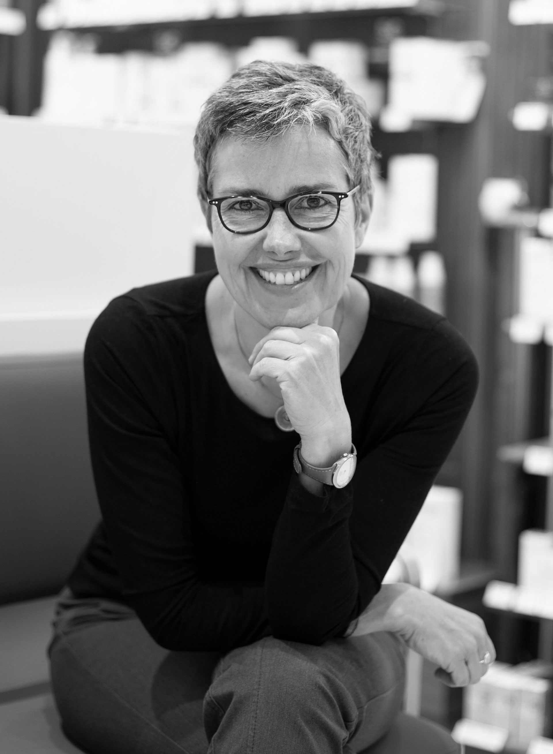 Frau mit Brille lächelt, das Kinn auf die Hand gestützt. Sie trägt ein schwarzes Hemd und sitzt drinnen.