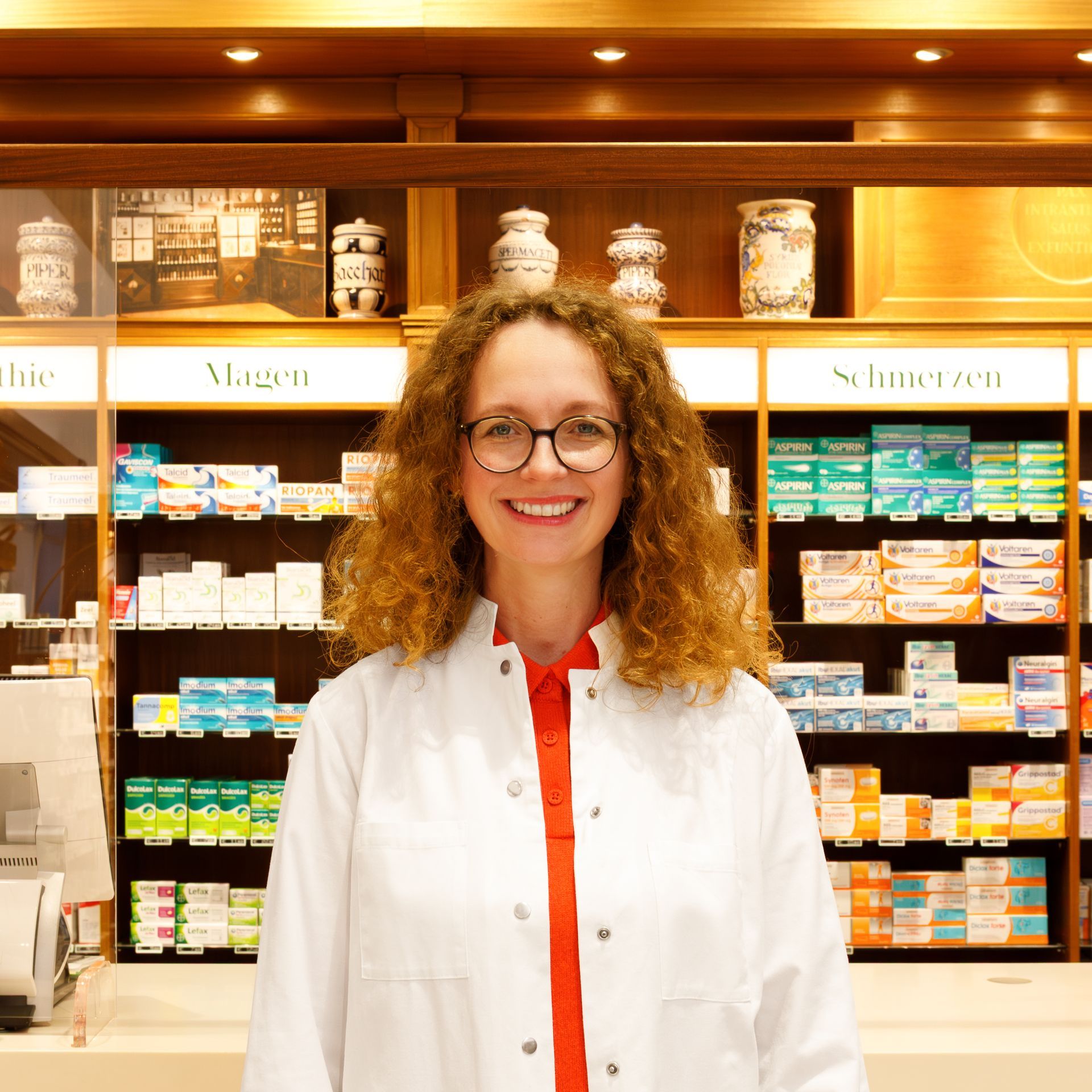 Eine Frau im Laborkittel lächelt in einer Apotheke; im Hintergrund sind Regale mit Medikamenten zu sehen.