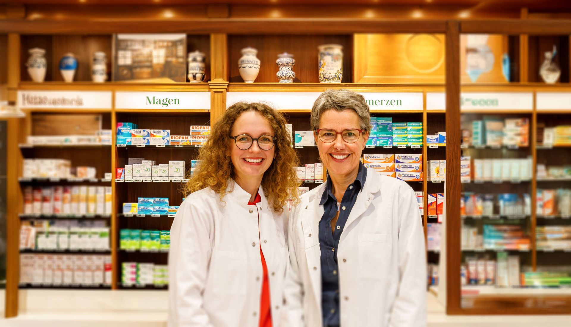 Zwei Personen in Laborkitteln lächeln in einer Apotheke mit Regalen voller Produkte.
