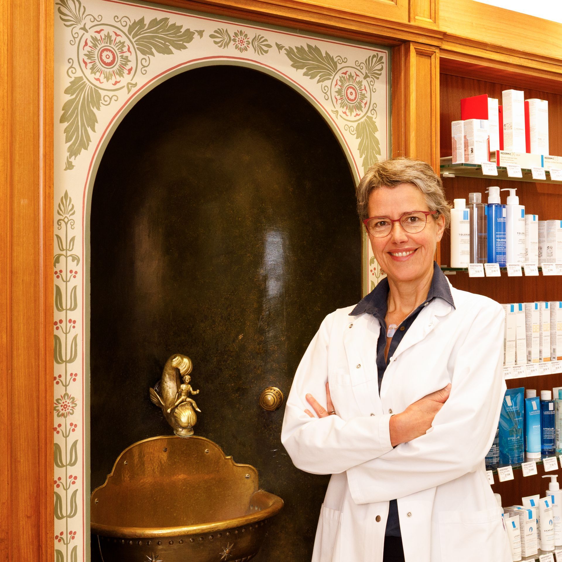Eine Frau im weißen Kittel steht in einer Apotheke. Sie hat die Arme verschränkt und lächelt. 