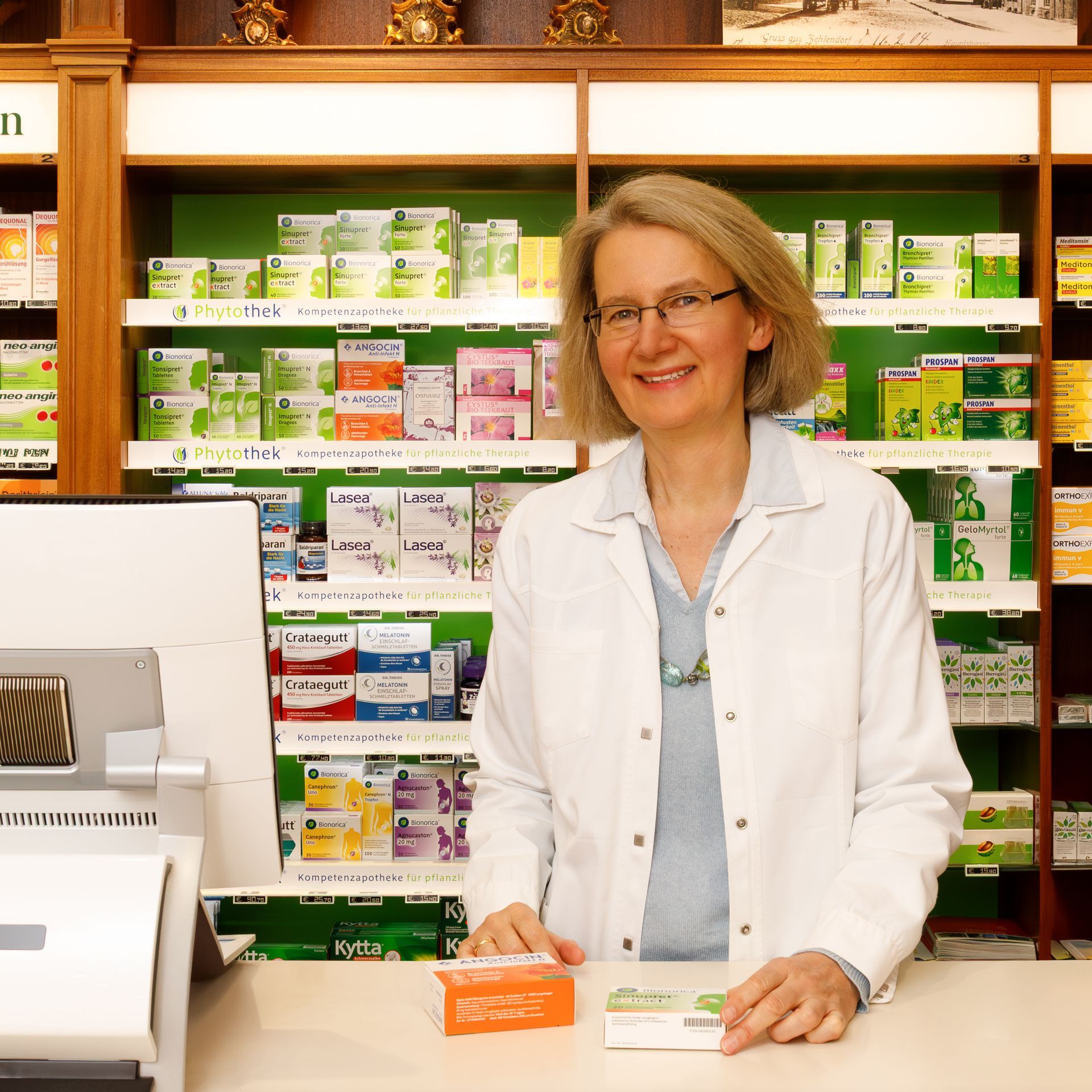 Ein Apotheker im weißen Kittel steht lächelnd am Apothekentresen. Dahinter stehen in Regalen Produkte in Kartons.