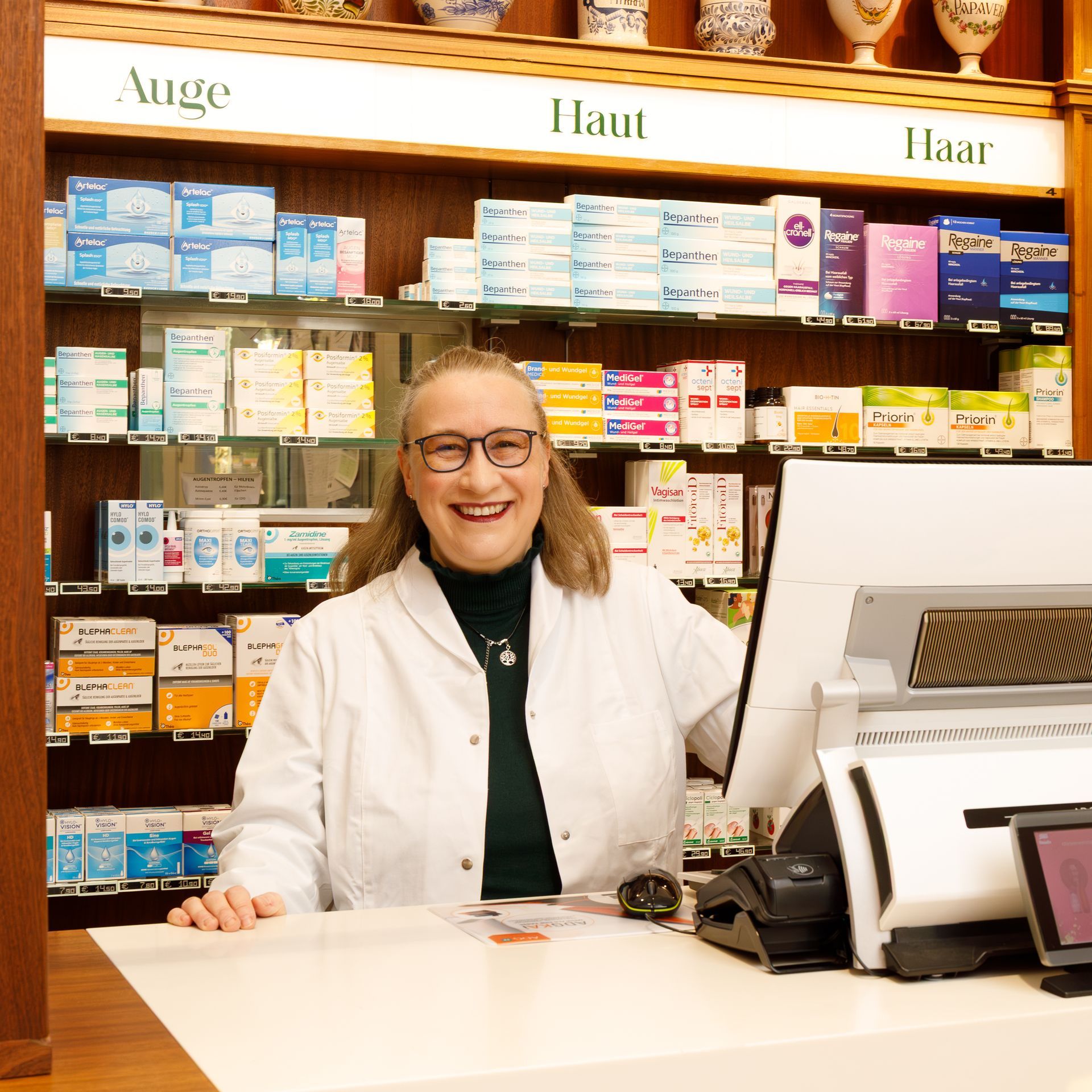 Hinter dem Apothekentresen lächelt ein Apotheker im weißen Kittel. In den Regalen stehen Medikamente.