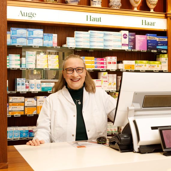 Hinter dem Apothekentresen lächelt ein Apotheker im weißen Kittel. In den Regalen stehen Medikamente.