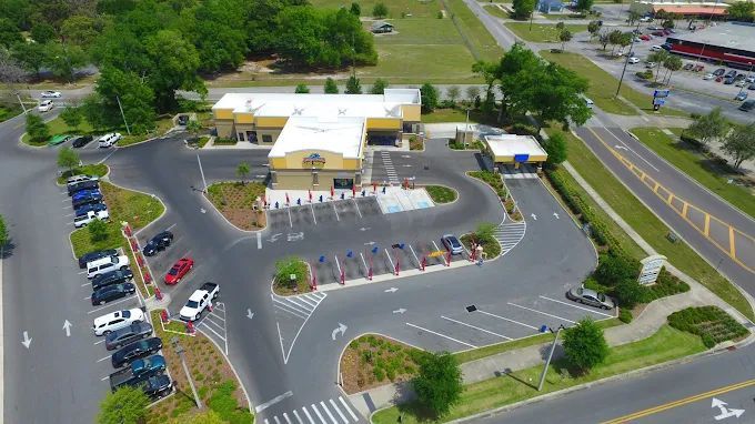 Aerial view of a car wash with cars parked. A winding driveway leads to the building and charging stations. Green trees and blue sky.