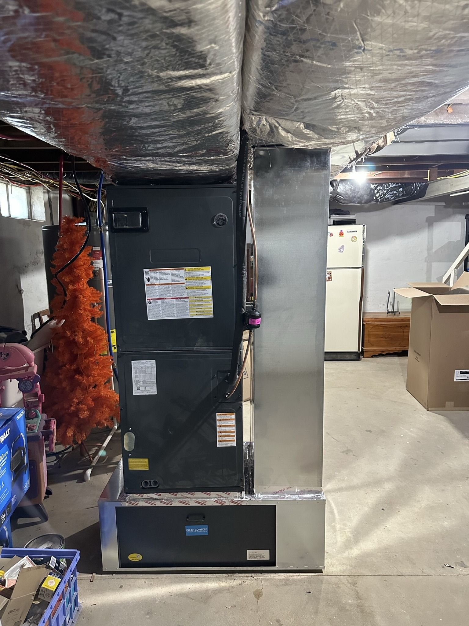 A grey HVAC furnace unit with shiny metal ductwork stands in a cluttered basement.