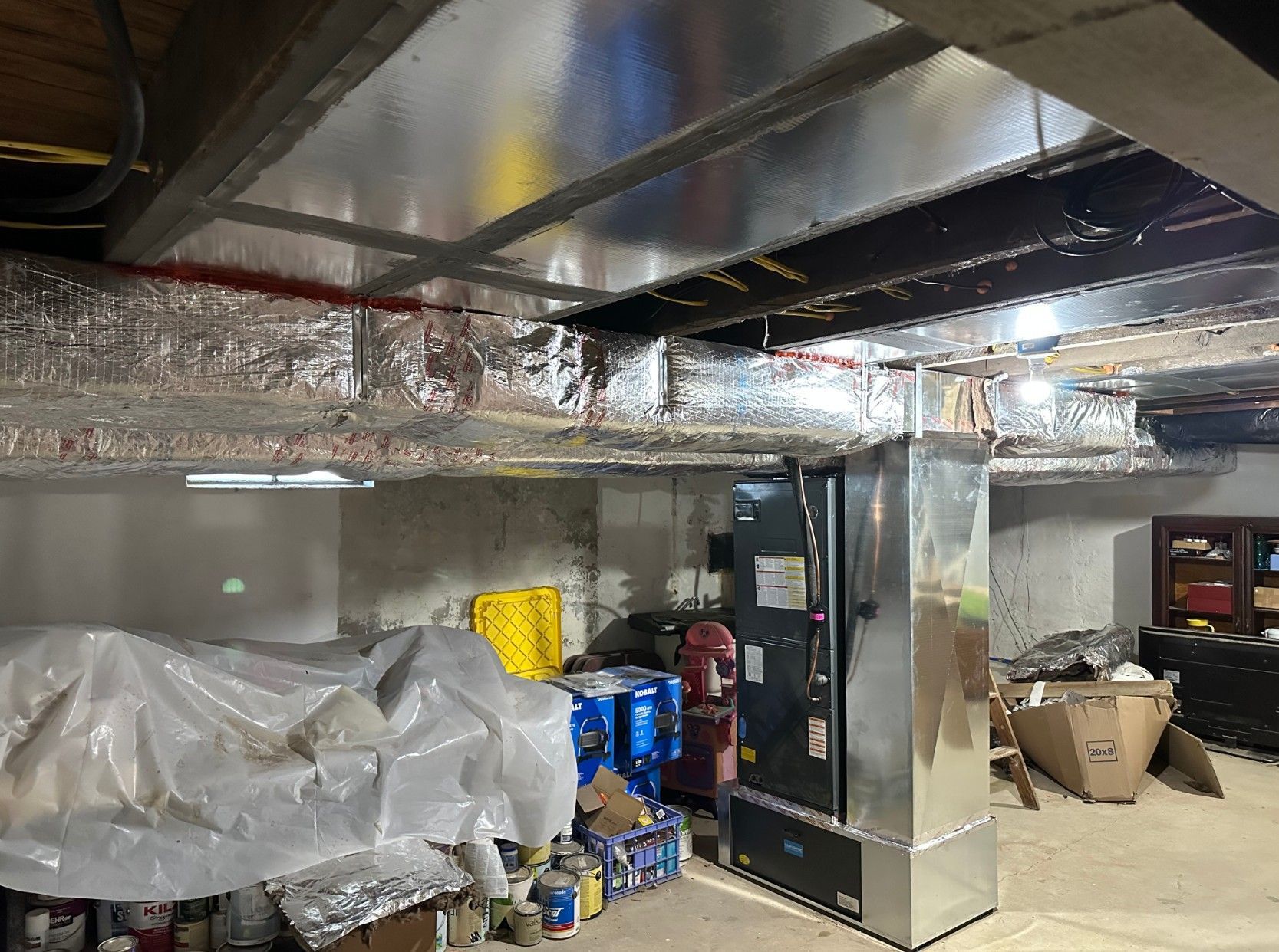 A basement utility area featuring a furnace, silver ventilation ductwork, and stored items under plastic sheeting.