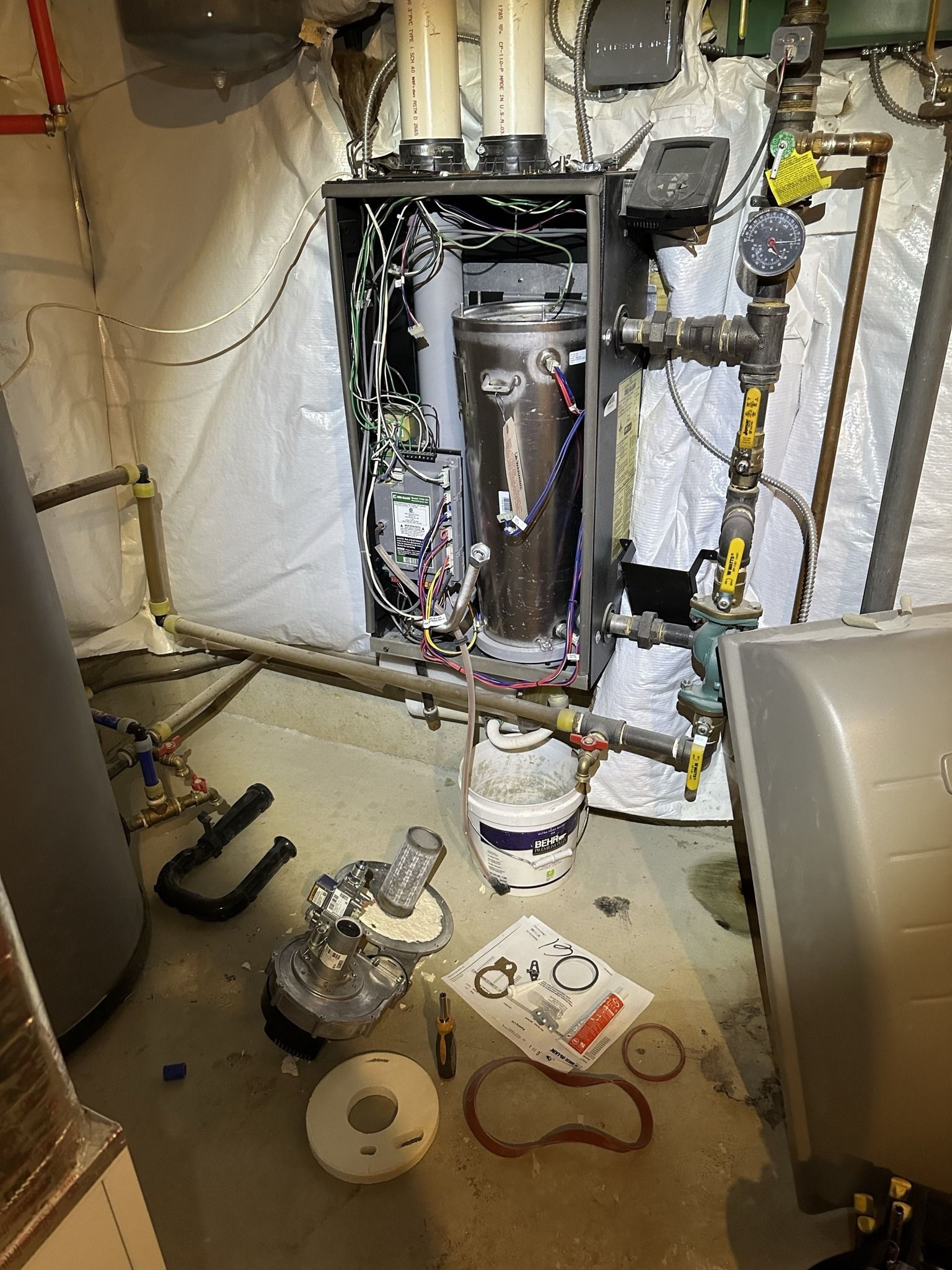 A disassembled high-efficiency boiler in a basement, with parts, a manual, and a white bucket placed on the floor.
