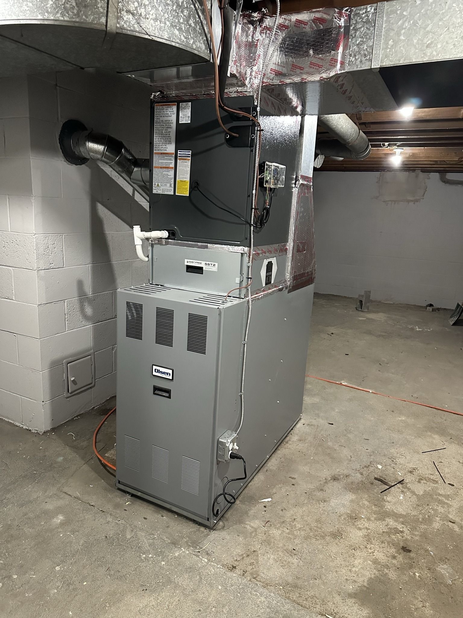 A grey HVAC furnace unit sits in a basement with concrete block walls, a concrete floor, and exposed ceiling joists.