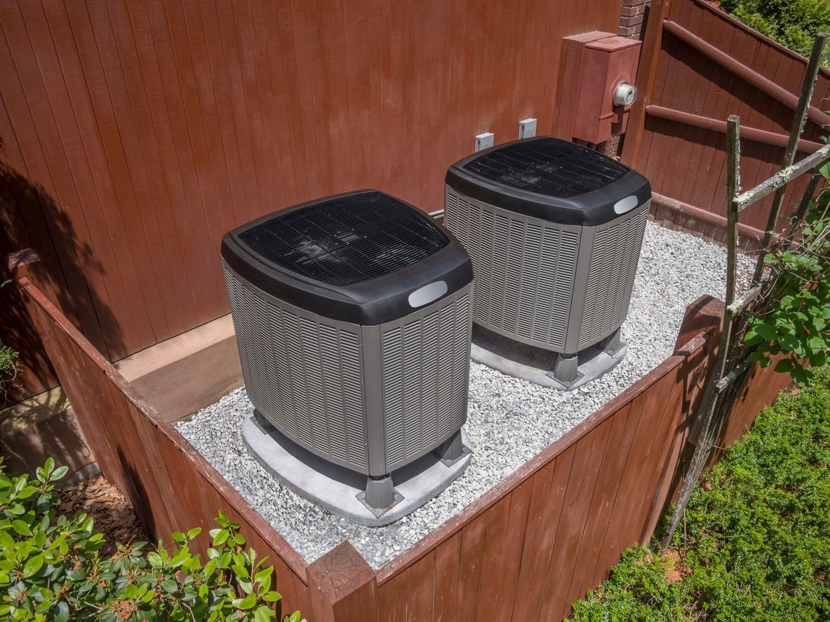 Two gray air conditioning units sit on a gravel bed next to a wooden wall.