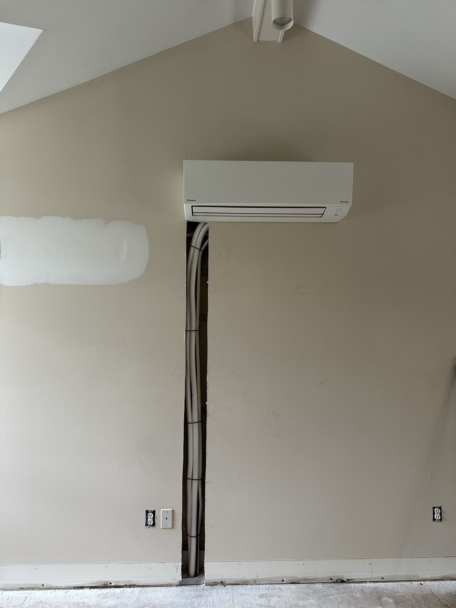 A ductless mini-split unit mounted on a tan wall with exposed refrigerant lines running down an open channel in the drywall.