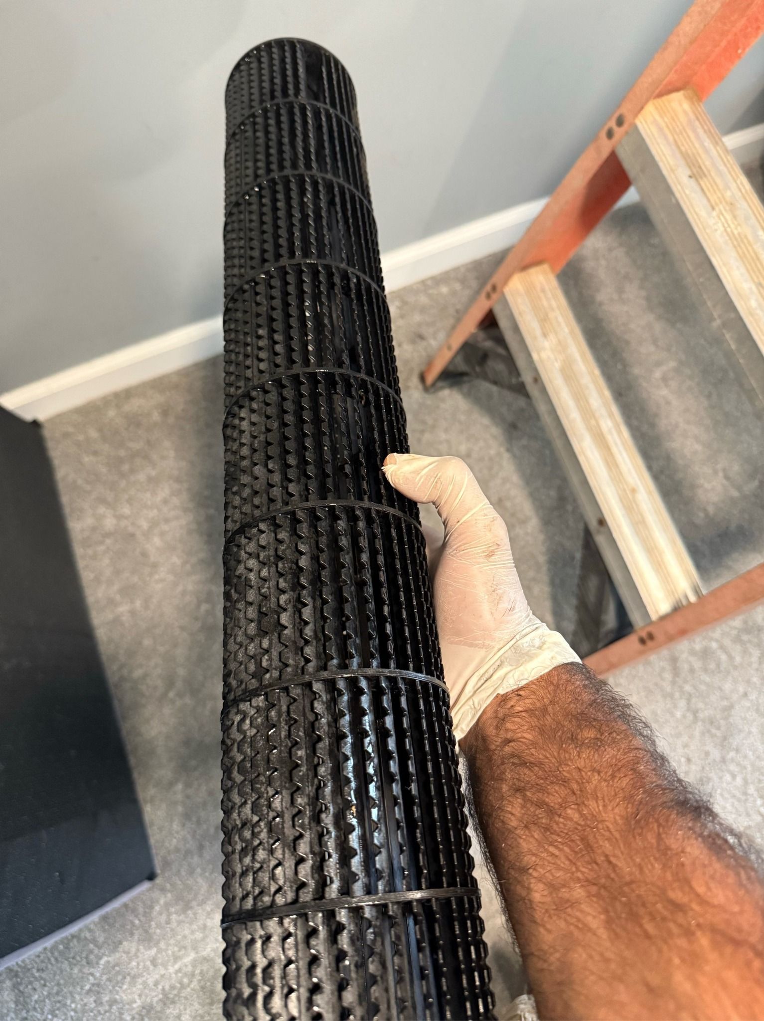 A person wearing a glove holds a long, black cylindrical HVAC blower wheel indoors next to a wooden stepladder.