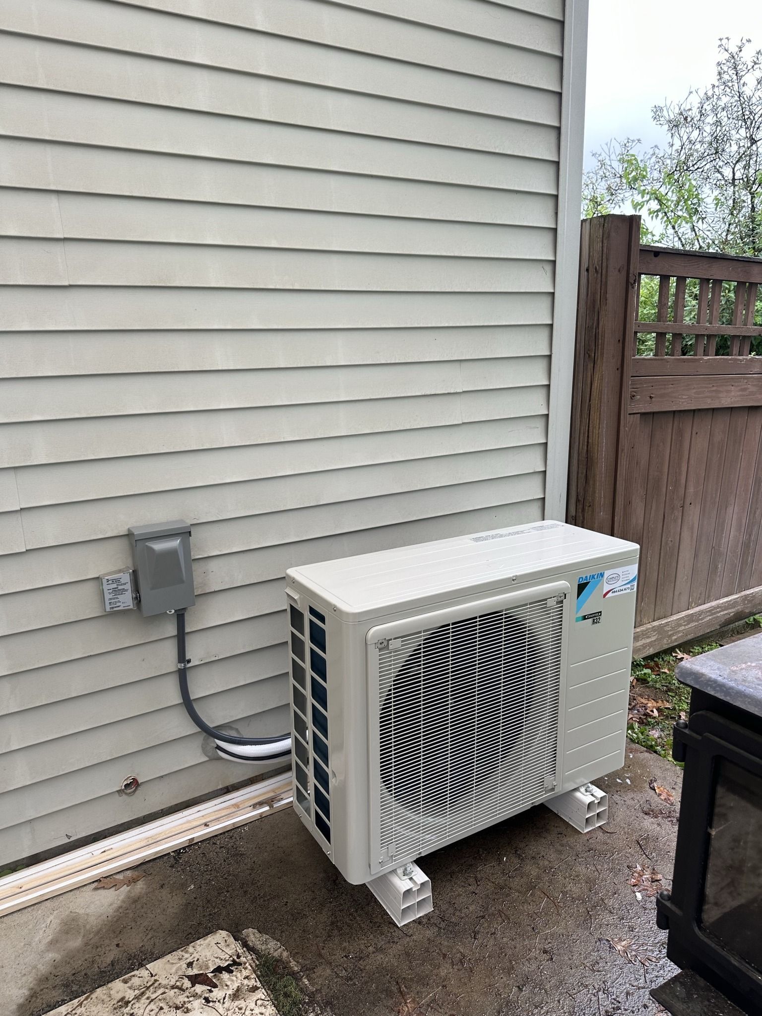A beige Daikin heat pump outdoor unit sits on a concrete patio against light-colored vinyl siding near a wooden fence.
