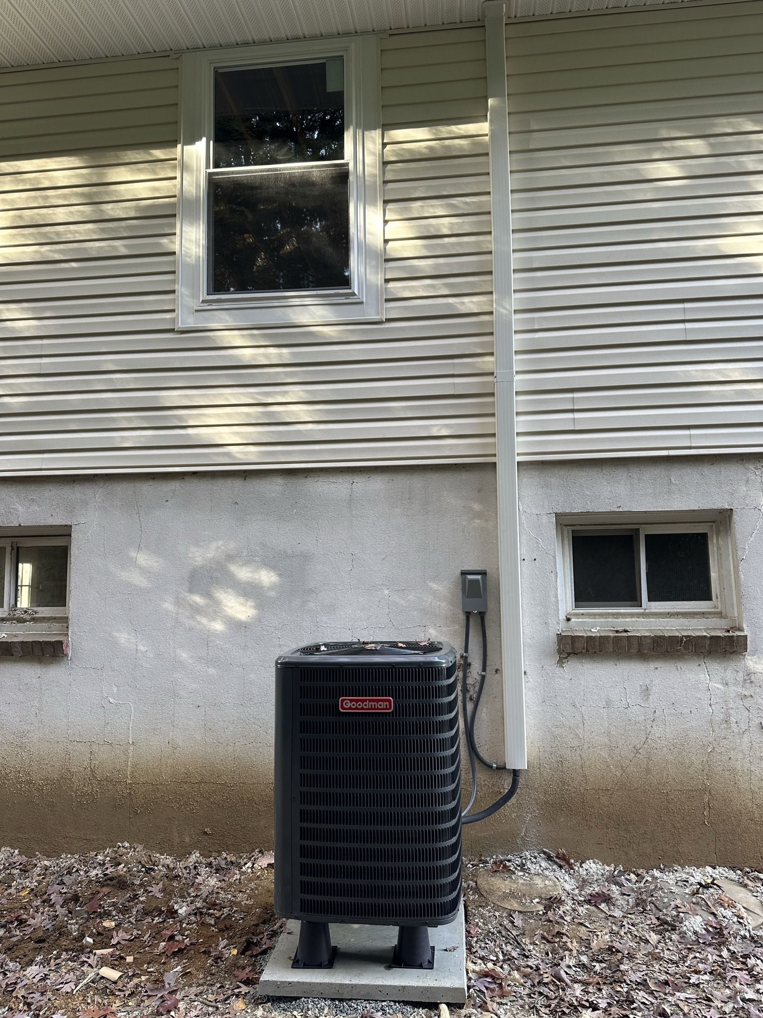 A black HVAC unit sits on a concrete pad outside a house with beige siding and a light-colored foundation.