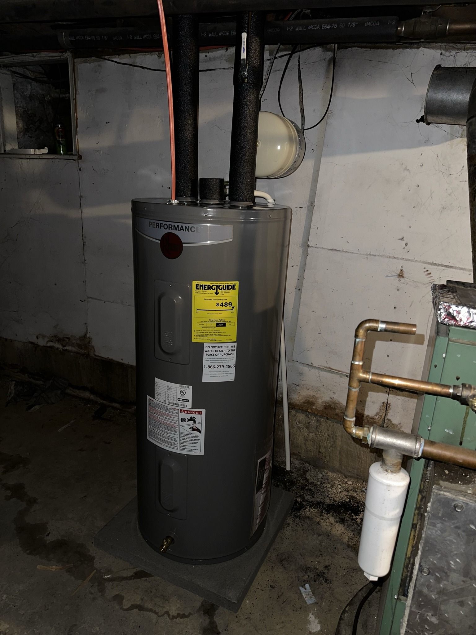 A grey residential electric water heater stands on a base in a concrete basement near plumbing pipes.
