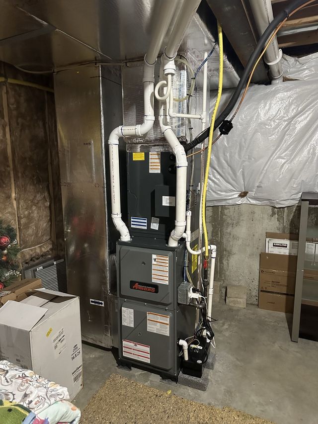 A gray furnace unit with white PVC intake and exhaust pipes standing in a basement corner near a concrete wall.