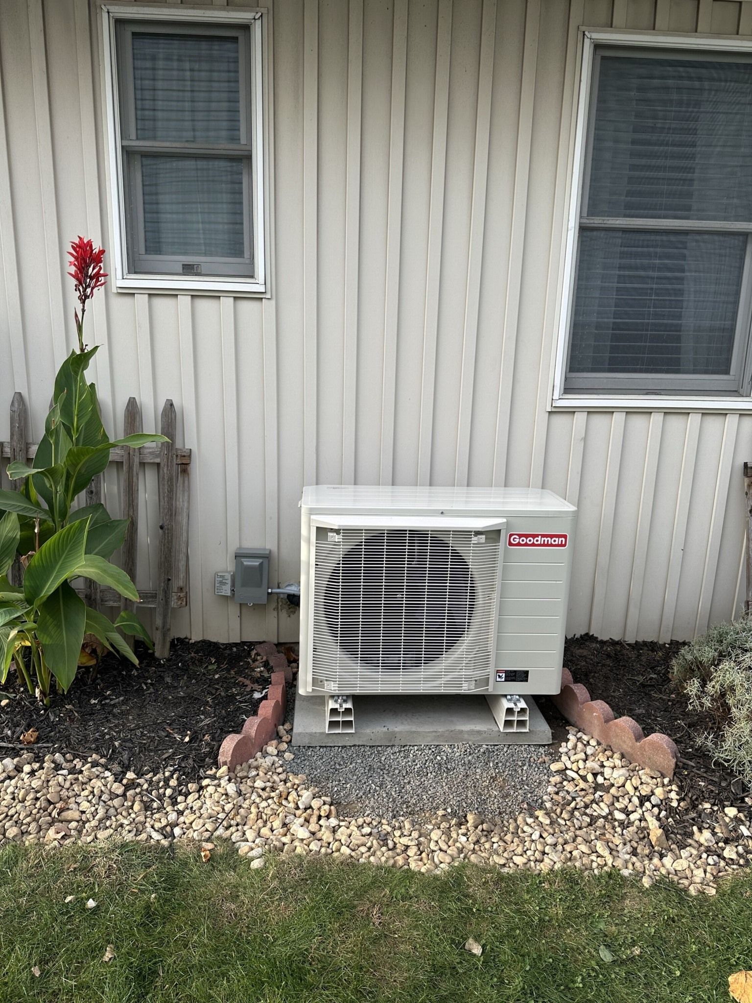 A light-colored outdoor air conditioning unit sits on concrete blocks against the side of a house with light siding.