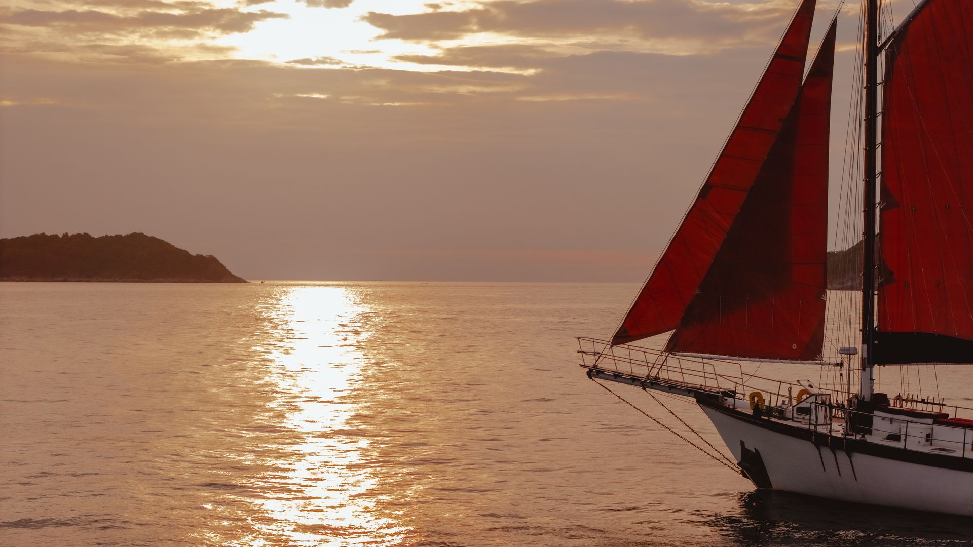 A sailboat - schooner - with red sails is in the ocean at sunset