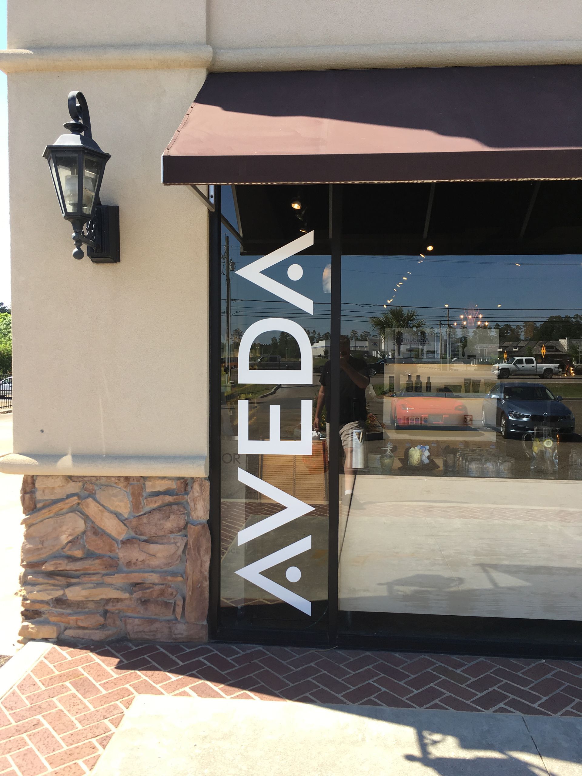 A storefront entrance with the white, vertical Aveda logo on the glass door, under a brown awning on a brick-accented building.