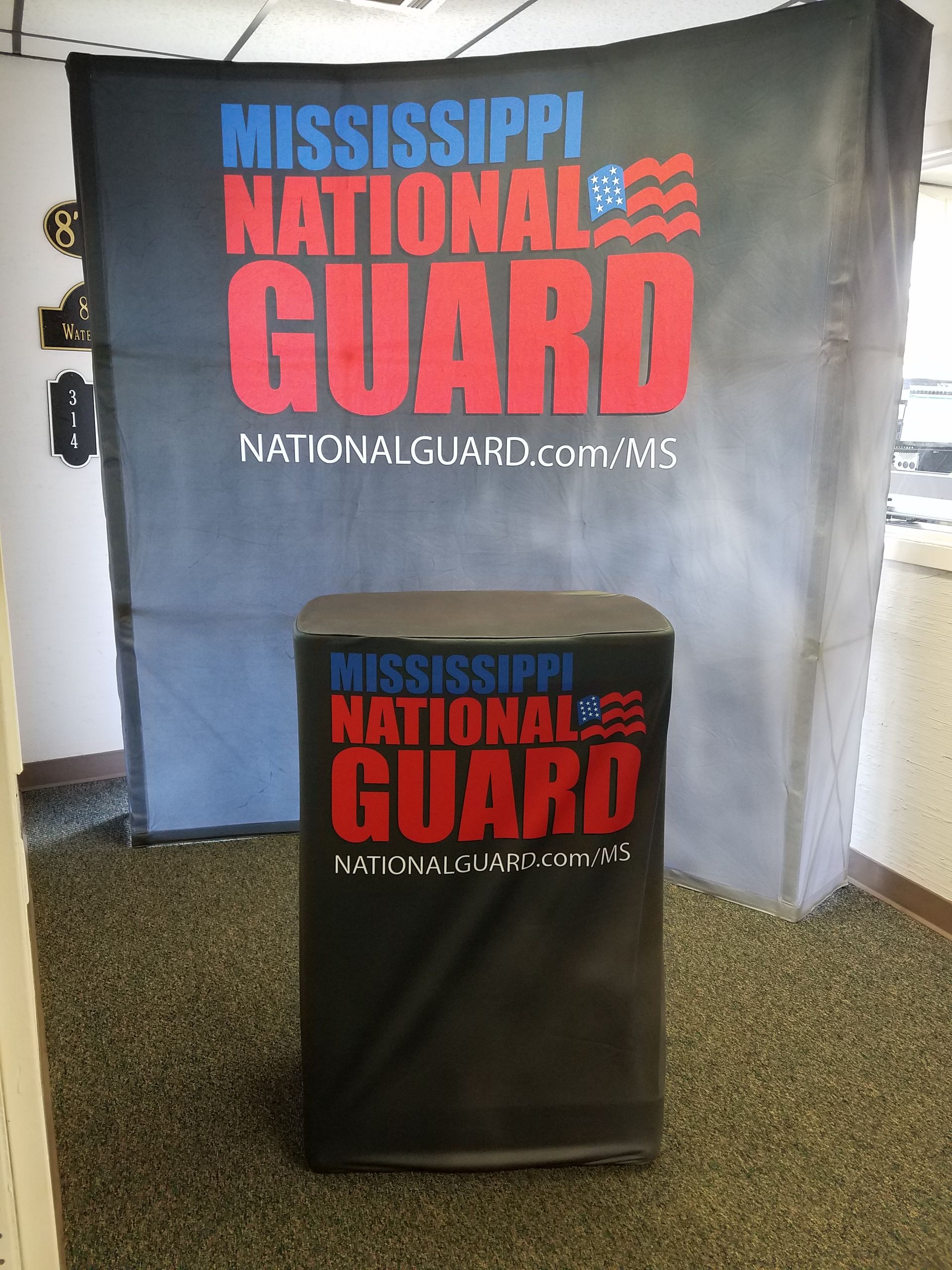 A Mississippi National Guard promotional display booth with a large backdrop and matching podium on a carpeted floor.