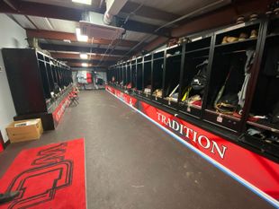 A sports locker room with dark stalls, a red "TRADITION" sign, and a large red team rug on a dark floor.